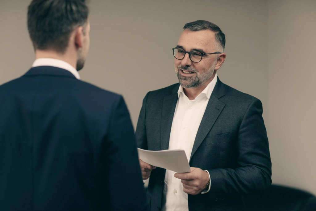 Smiling man in suit and a paper deck in hand talks to another man in suit