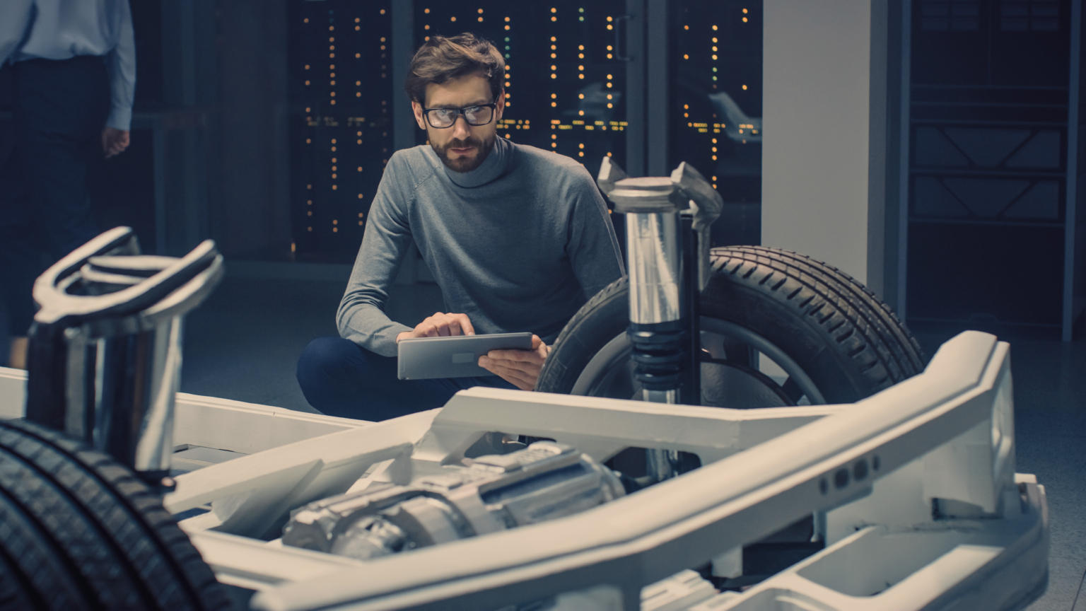 Automobile Design Engineer Sitting Beside Hybrid Electric Car Chassis Platform Prototype, Using Tablet Computer for Design Enhancement. Facility with Vehicle Frame with Suspension, Wheels, Engine and Battery