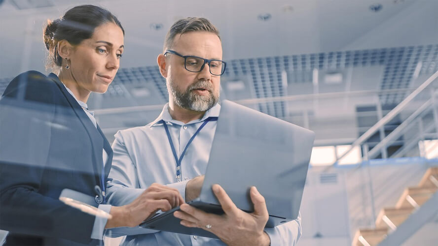 Business man is showing something to a business woman on his laptop. Modern facility in the background. Low angle shot.