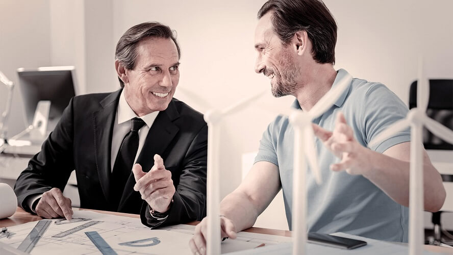 One business man is talking to a casual man. Both sitting on a desk and laughing while speaking about some engineering project work.